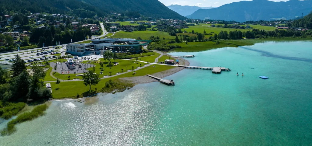 Im Hintergrund Berge und grüne Wiesen, davor ein Gebäudekomplex mit Steg. Im Vordergrund ein großer See mit azurblauem Wasser